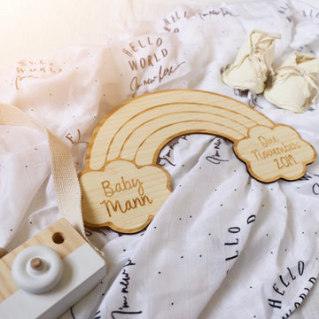 The Personalised Rainbow Announcement Plaque, crafted from eco-friendly bamboo and engraved with your custom details, placed on a white blanket with baby accessories.