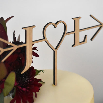 An arrow shaped cake topper in dark bamboo, with a heart and the letters J and L. Sitting on a white cake with floral arrangement. 
