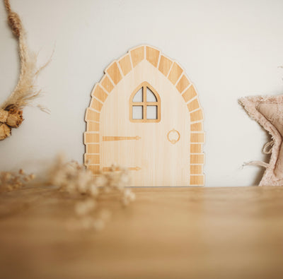 A light bamboo arch shaped fairy door against a wall. It has an etched window, hinges and door handle. Sitting amongst decorative florals. 