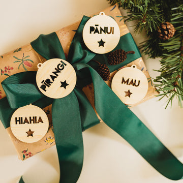 A gift wrapped in floral paper with a green ribbon is decorated with Te Reo - Want, Need, Wear, Read Tags (Unpersonalised), each featuring Māori words and star-shaped cutouts. Pine sprigs are placed nearby.