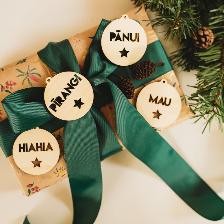 A gift wrapped in floral paper with a green ribbon is decorated with Te Reo - Want, Need, Wear, Read Tags (Unpersonalised), each featuring Māori words and star-shaped cutouts. Pine sprigs are placed nearby.