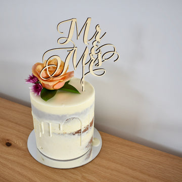 A light bamboo cake topper saying Mr and Mrs, sitting on a white drip cake with a floral arrangement.