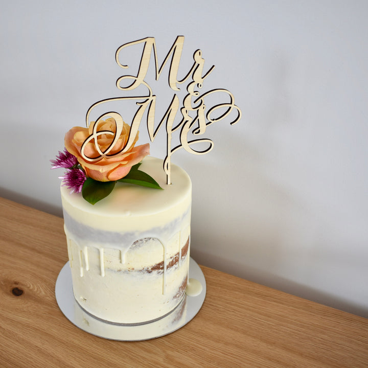 A light bamboo cake topper saying Mr and Mrs, sitting on a white drip cake with a floral arrangement.