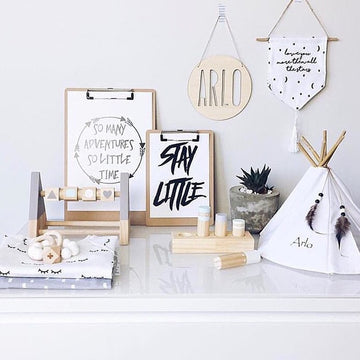 A minimalist nursery display features wooden toys, a small tipi labeled Arlo, a potted succulent, two quote prints on clipboards, a Personalised Natural Names Plaque, and a decorative wall hanging against a white backdrop.