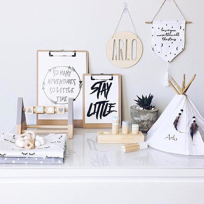 A minimalist nursery display features wooden toys, a small tipi labeled Arlo, a potted succulent, two quote prints on clipboards, a Personalised Natural Names Plaque, and a decorative wall hanging against a white backdrop.
