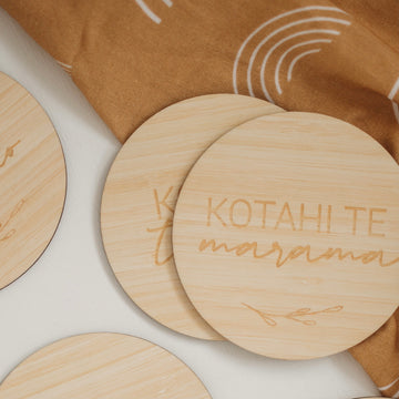 A round light bamboo set of baby milestones, lying on a brown baby blanket with white rainbows. The two visible sign says kotahi te marama (one month).