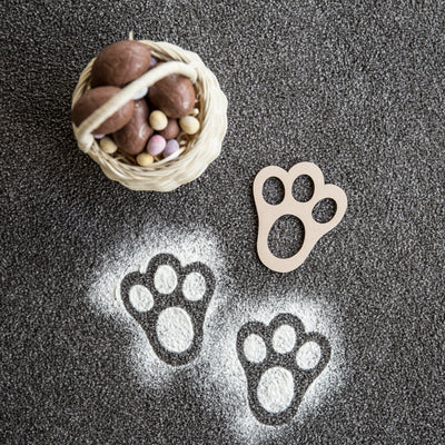 An MDF Easter bunny stencil, lying on the ground next to an Easter basket full of Easter eggs, and two bunny foot prints made with flour. 