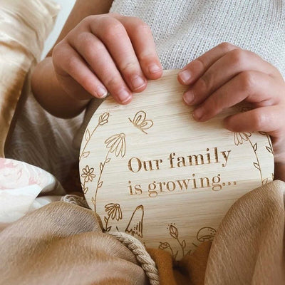 Little hands holding a round bamboo saying Our family is growing... with a floral border by Chloe Mytton.