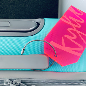 A turquoise suitcase with a grey handle features the Personalised Luggage Tag, attached by a metal loop. The tag is pink and displays the name Kylie in large cursive letters.