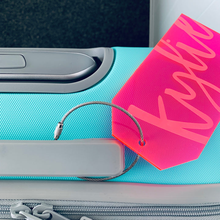 A turquoise suitcase with a grey handle features the Personalised Luggage Tag, attached by a metal loop. The tag is pink and displays the name Kylie in large cursive letters.