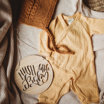 A round light bamboo plaque saying hush little baby, sitting with a yellow baby outfit and white blanket. 