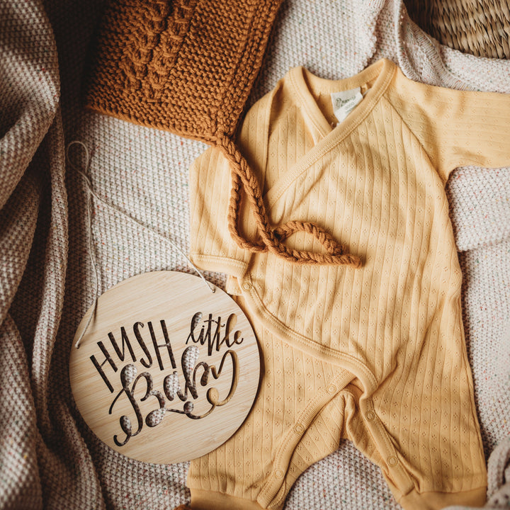 A round light bamboo plaque saying hush little baby, sitting with a yellow baby outfit and white blanket. 