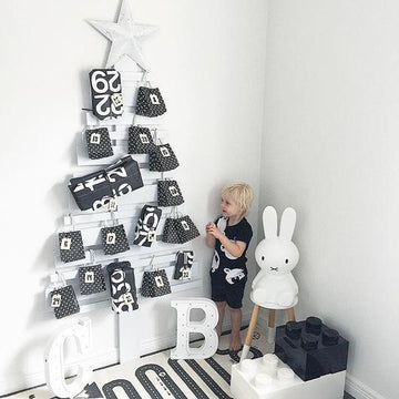 A young child stands beside a minimalist white Christmas tree decorated with the Advent Calendar Tags Set, with a large bunny lamp and oversized LEGO blocks.