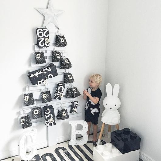 A young child stands beside a minimalist white Christmas tree decorated with the Advent Calendar Tags Set, with a large bunny lamp and oversized LEGO blocks.