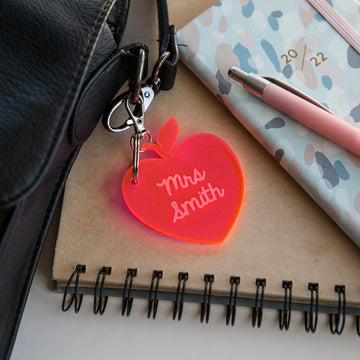 A Teacher Key Ring shaped like a red apple and engraved with "Mrs Smith" sits on a spiral notebook beside a pink pen, patterned planner, and black bag strap—a unique personalized teacher gift.
