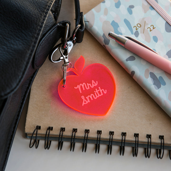 A Teacher Key Ring shaped like a red apple and engraved with "Mrs Smith" sits on a spiral notebook beside a pink pen, patterned planner, and black bag strap—a unique personalized teacher gift.