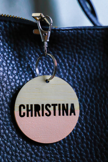 A round light bamboo keyring, half painted coral, saying Christina. Attached to a black leather bag. 