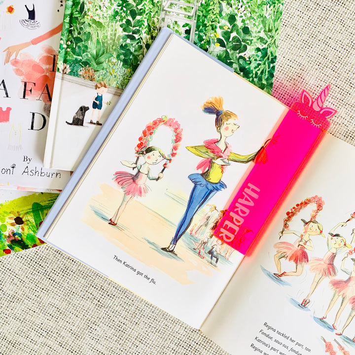 A neon pink see through acrylic bookmark with a unicorns head at the end, and the name Harper etched on, sitting atop a children&