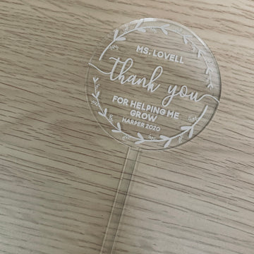 The Personalised Wreath Teacher Planters feature a round acrylic plaque with white floral designs and an engraved message: Ms. Lovell, thank you for helping me grow, Harper 2020. This teacher gift rests on a light wood base.