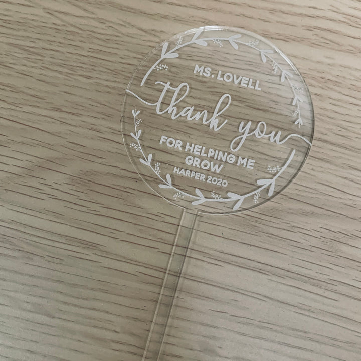 The Personalised Wreath Teacher Planters feature a round acrylic plaque with white floral designs and an engraved message: Ms. Lovell, thank you for helping me grow, Harper 2020. This teacher gift rests on a light wood base.