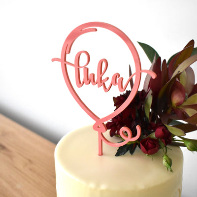 A pink acrylic balloon shaped cake topper, with the name Luka in linked writing inside. Sitting atop a white cake with floral arrangement. 