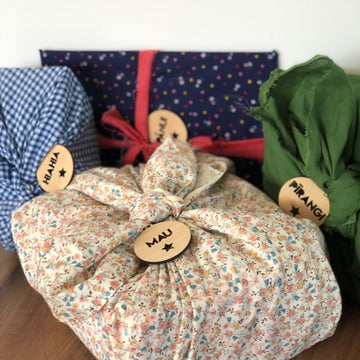 Four gifts wrapped in patterned fabric, each tied with cloth and labelled with Te Reo - Want, Need, Wear, Read Tags (Unpersonalised). The gifts are displayed on a wooden surface against a plain background.
