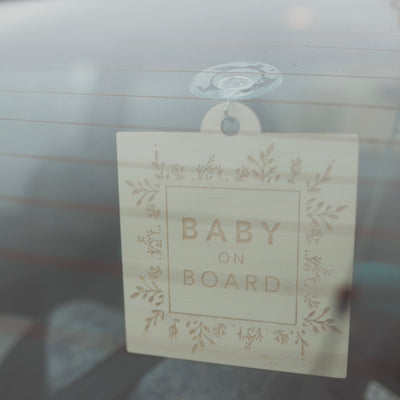 A light bamboo sign saying Baby on Board, with a border of engraved laurels and florals. Hanging from a car window.