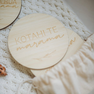 A round light bamboo set of baby milestones in a white calico bag, lying on a baby blanket with a rattle. The two visible sign says kotahi te marama (one month).