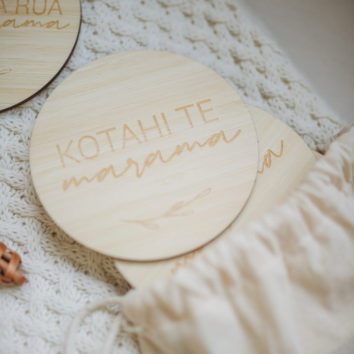 A round light bamboo set of baby milestones in a white calico bag, lying on a baby blanket with a rattle. The two visible sign says kotahi te marama (one month).