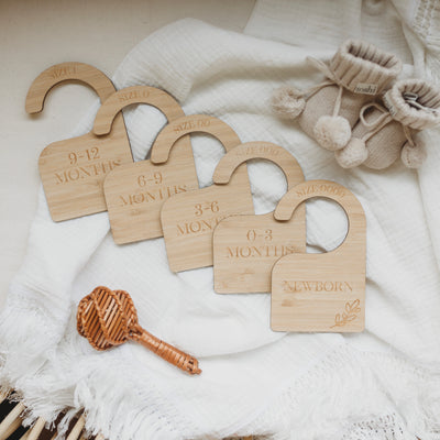 Five bamboo wardrobe dividers. On the top are the sizes 0000, 000, 00, 0 and 1 and on the bottom is a laurel engraved and the months. Newborn, 0-3 months, 3-6 months, 6-9 months, 9-12 months. Sitting atop a white shawl with a rattle and booties. 