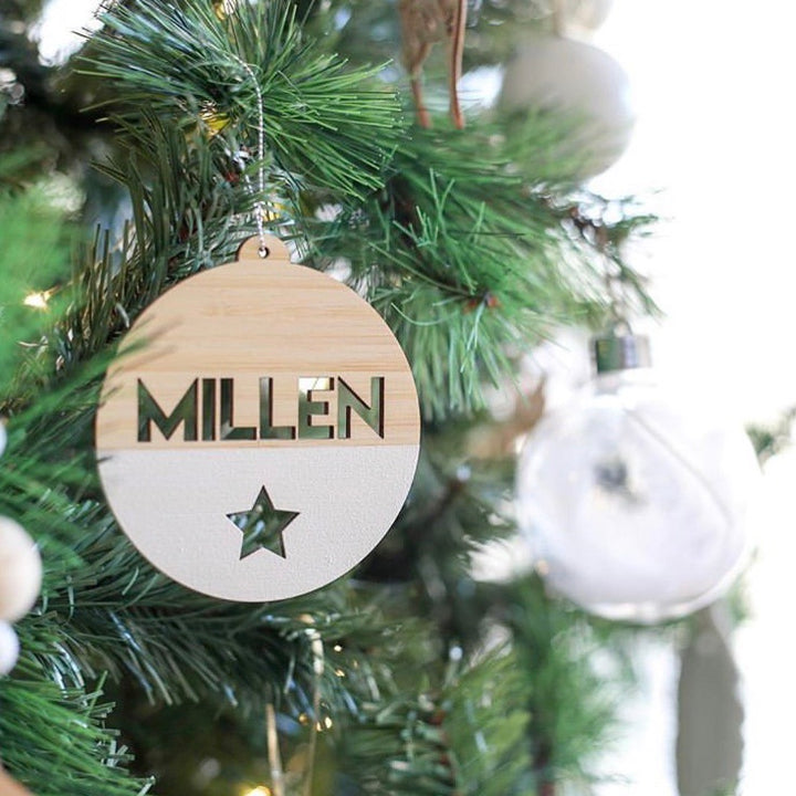 The Original Personalised Christmas Bauble hangs on a green Christmas tree with "MILLEN" and a cut-out star, adding charm to your family tradition while another clear round bauble sparkles in the background.