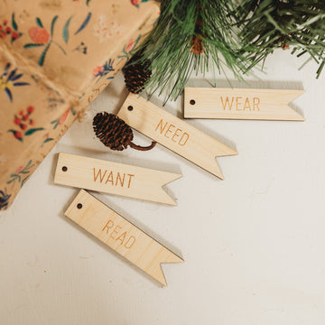 Four light bamboo tags in the shape of a flag saying want, need, wear and red. Sitting next to a brown paper wrapped present and pine cones. 