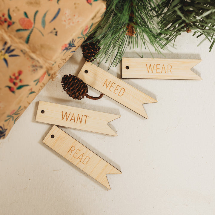 Four light bamboo tags in the shape of a flag saying want, need, wear and red. Sitting next to a brown paper wrapped present and pine cones. 