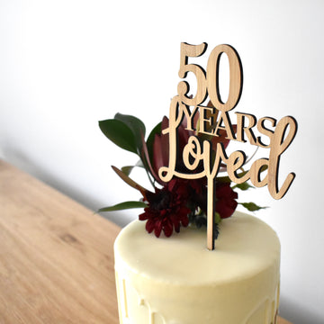 A white frosted cake with dark red flowers and green leaves is topped with a Years Loved Personalised Cake Topper that reads "50 Years Loved," displayed on a wooden table against a plain background.