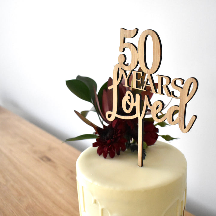 A white frosted cake with dark red flowers and green leaves is topped with a Years Loved Personalised Cake Topper that reads "50 Years Loved," displayed on a wooden table against a plain background.