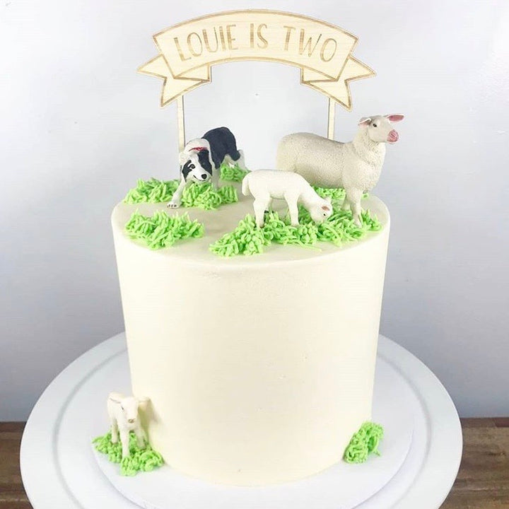 A bamboo banner cake topper saying Louie is Two, atop a white farmyard theme cake.
