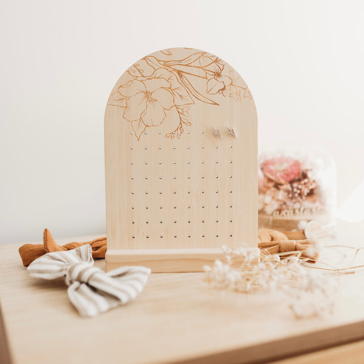 An arch shaped bamboo earring holder with a floral design etched on the top. Surrounded by hairbows.