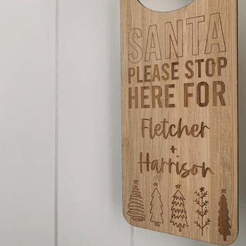 A wooden Santa Stop Here Door Hanger reads SANTA PLEASE STOP HERE FOR Fletcher + Harrison with engraved Christmas trees below, hanging on a white paneled wall—a charming, personalised Christmas tradition.