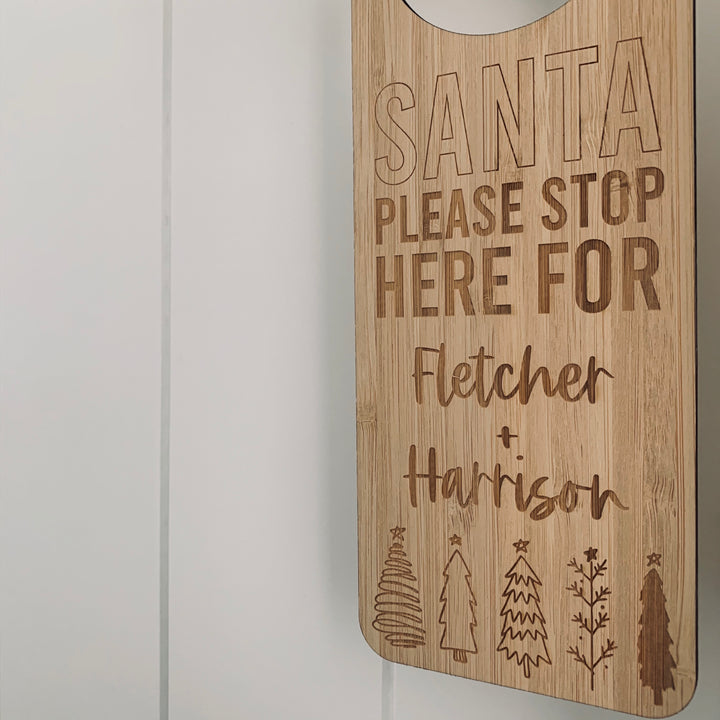 A wooden Santa Stop Here Door Hanger reads SANTA PLEASE STOP HERE FOR Fletcher + Harrison with engraved Christmas trees below, hanging on a white paneled wall—a charming, personalised Christmas tradition.