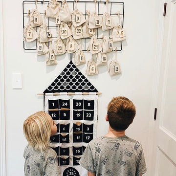 Two children curiously examine the Advent Calendar Tags Set displayed on a wall-mounted bamboo advent calendar with numbered cloth bags, alongside a fabric calendar in black, white, and neutral tones.