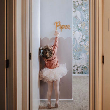A young girl in a tutu reaches up toward a gold Lovely Lettering Handlettered Wall Name on a pastel wall, with a hallway and unicorn-patterned wallpaper in the background.