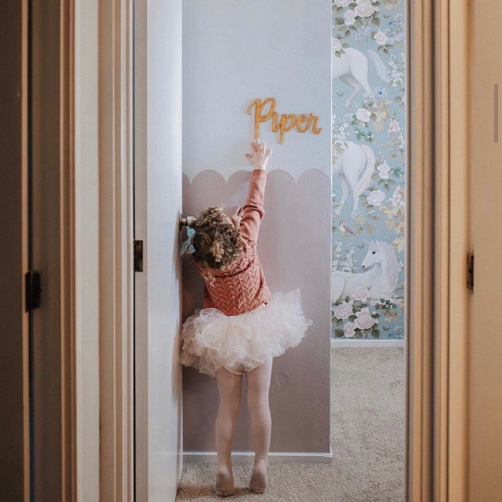 A young girl in a tutu reaches up toward a gold Lovely Lettering Handlettered Wall Name on a pastel wall, with a hallway and unicorn-patterned wallpaper in the background.