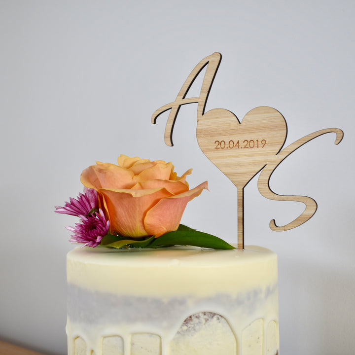 A dark bamboo cake topper in the shape of heart with the letters A and S on either side. Etched inside the heart is 20.04.2019. Sitting on a white cake with floral arrangement.