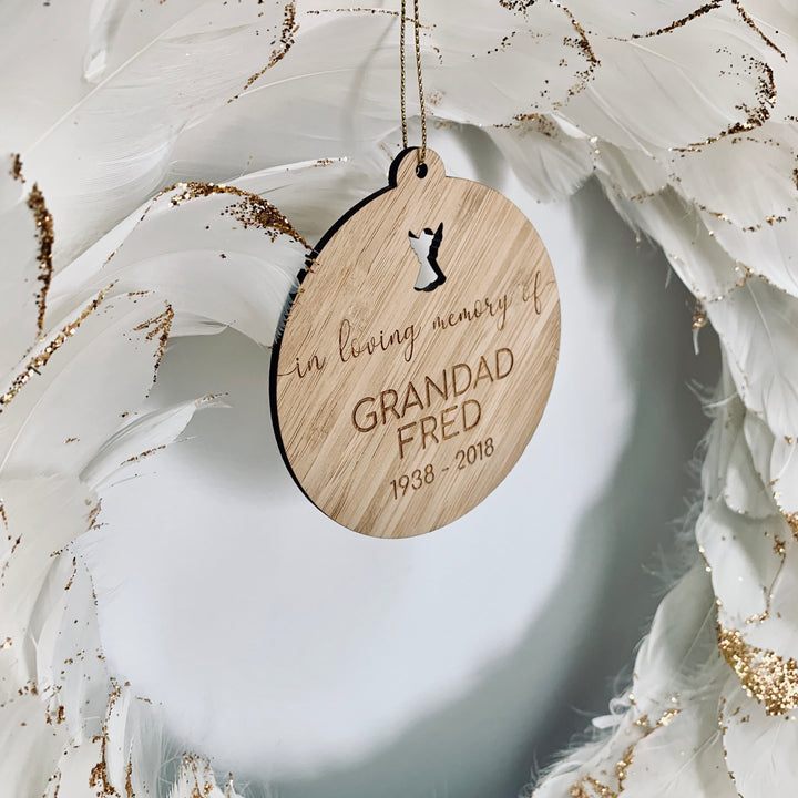 The In Loving Memory Christmas Bauble, engraved with “In loving memory of Grandad Fred 1938 - 2018,” hangs on a white feathered wreath with gold glitter, creating a touching memorial ornament.
