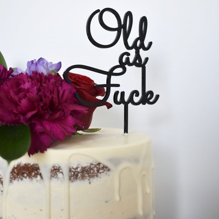 A white-frosted cake with a white chocolate drip, decorated with purple and red flowers, is topped with the Old As F... Cake Topper (other colours available) for a humorous birthday finish.