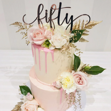 A two-tiered cake with pink and white roses, green leaves, gold accents, and pink drip icing is topped with a Mirror Personalised Cake Topper reading fifty to celebrate a 50th milestone. The cake is displayed on a white tablecloth.