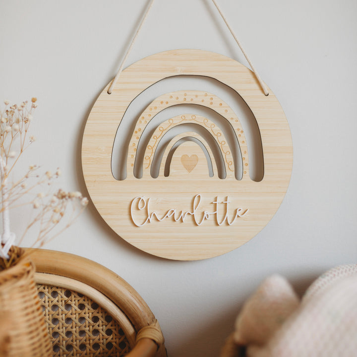 The Personalised Rainbow Layered Plaque features rainbow shapes, a heart, and the name Charlotte in cursive script. Displayed above a wicker chair with dried flowers on the left, it adds a charming touch to any room.