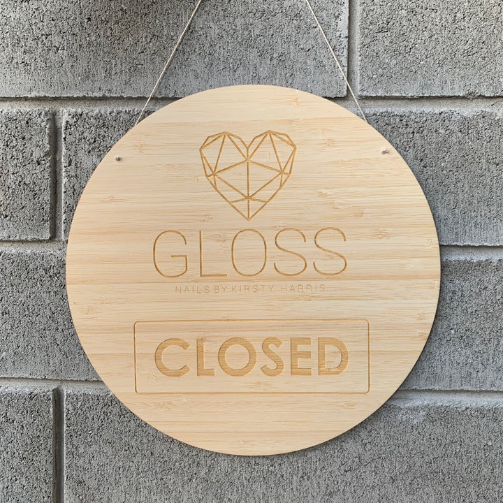 A light bamboo round sign saying closed, with Gloss Nails logo engraved. Hanging against a grey brick wall.