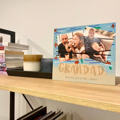 A rectangle MDF photo holder saying Grandad. Made with love by Indie + Harper. It has been drawn on by a child, and has two picture of two girls with their Grandad.  A piece of black elastic is holding the photos in place. 