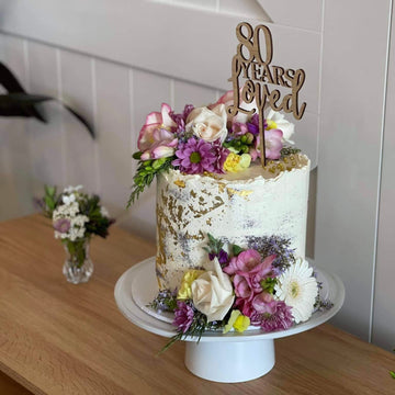 A white cake with gold accents and fresh flowers sits on a stand, topped with the Years Loved Personalised Cake Topper reading “80 years loved.” A matching floral arrangement is placed beside the cake on a wooden table.